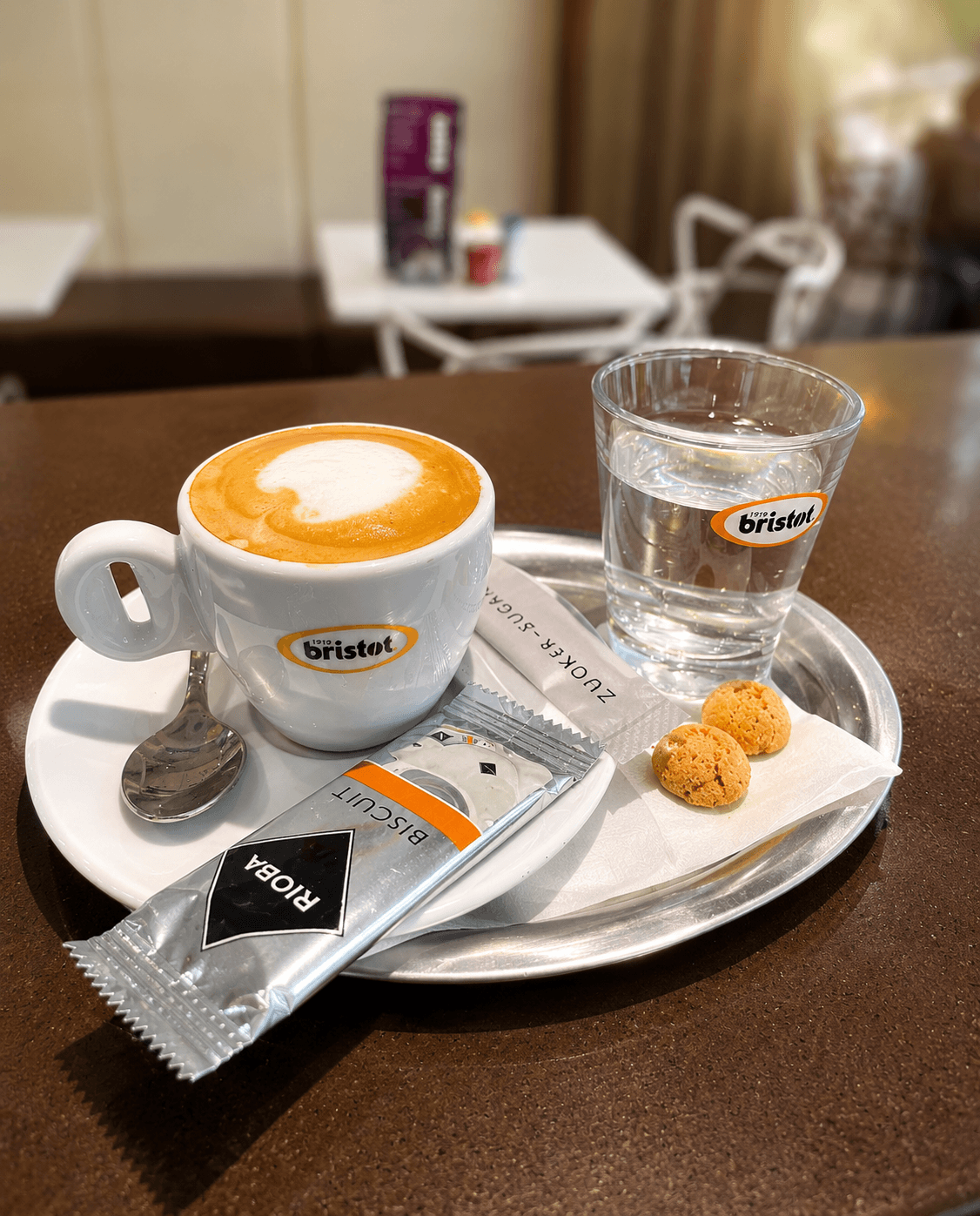 Cappuccino mit Milchschaum, ein Glas Wasser und Kekse auf einem silbernen Tablett im Café.