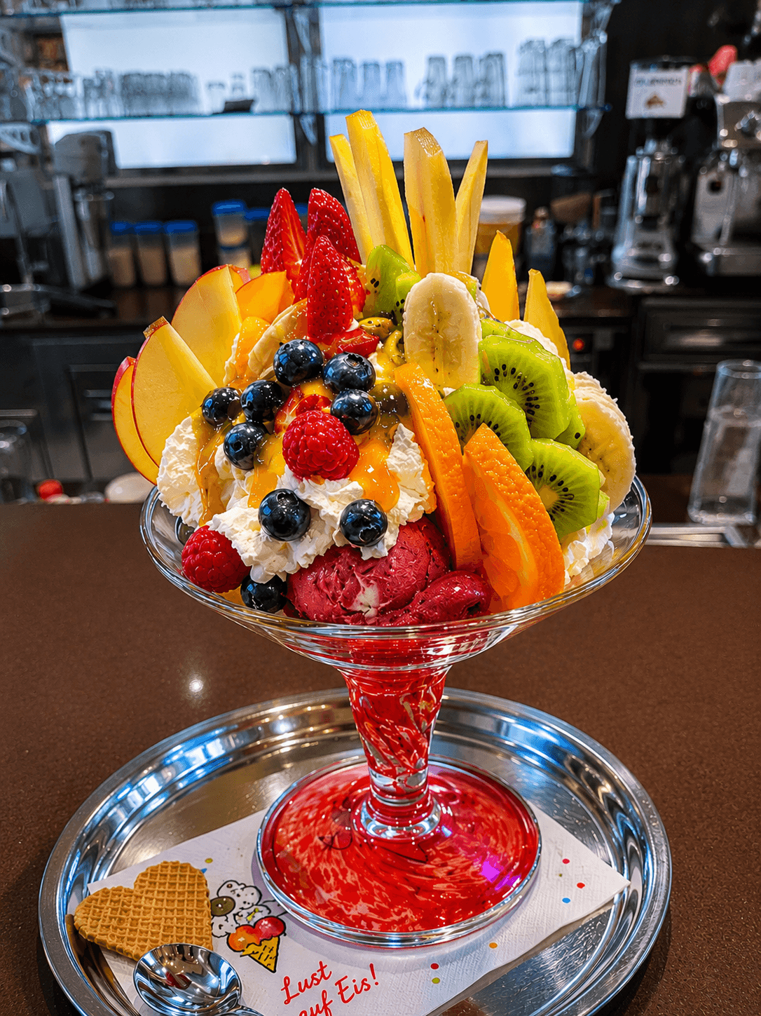 Farbenfroher Eisbecher mit frischen Früchten, Schlagsahne und rotem Sorbet in einer Glasschale auf Silbertablett.