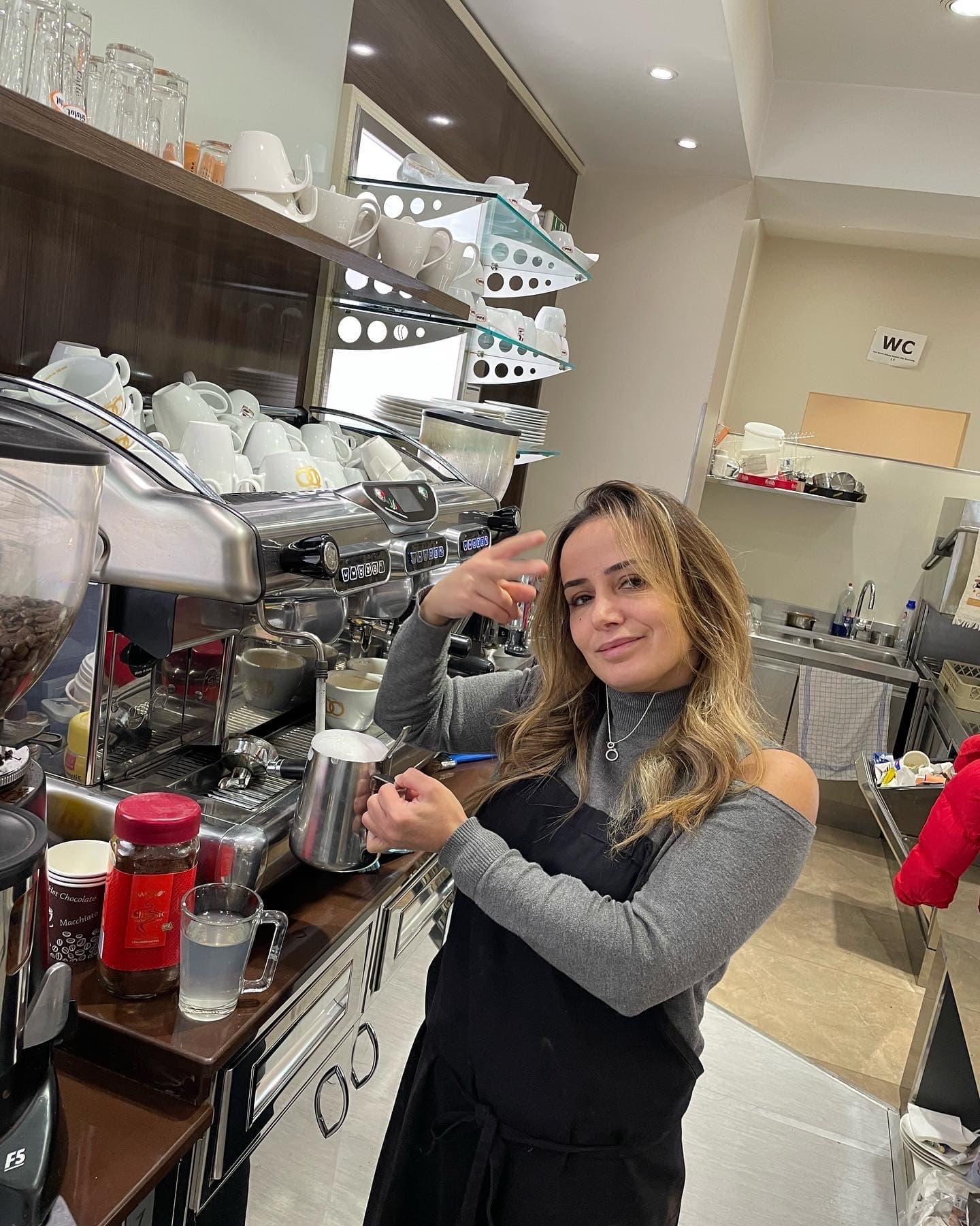 Lächelnde Barista mit Friedenszeichen posiert in schwarzer Schürze hinter einer professionellen Espressomaschine.