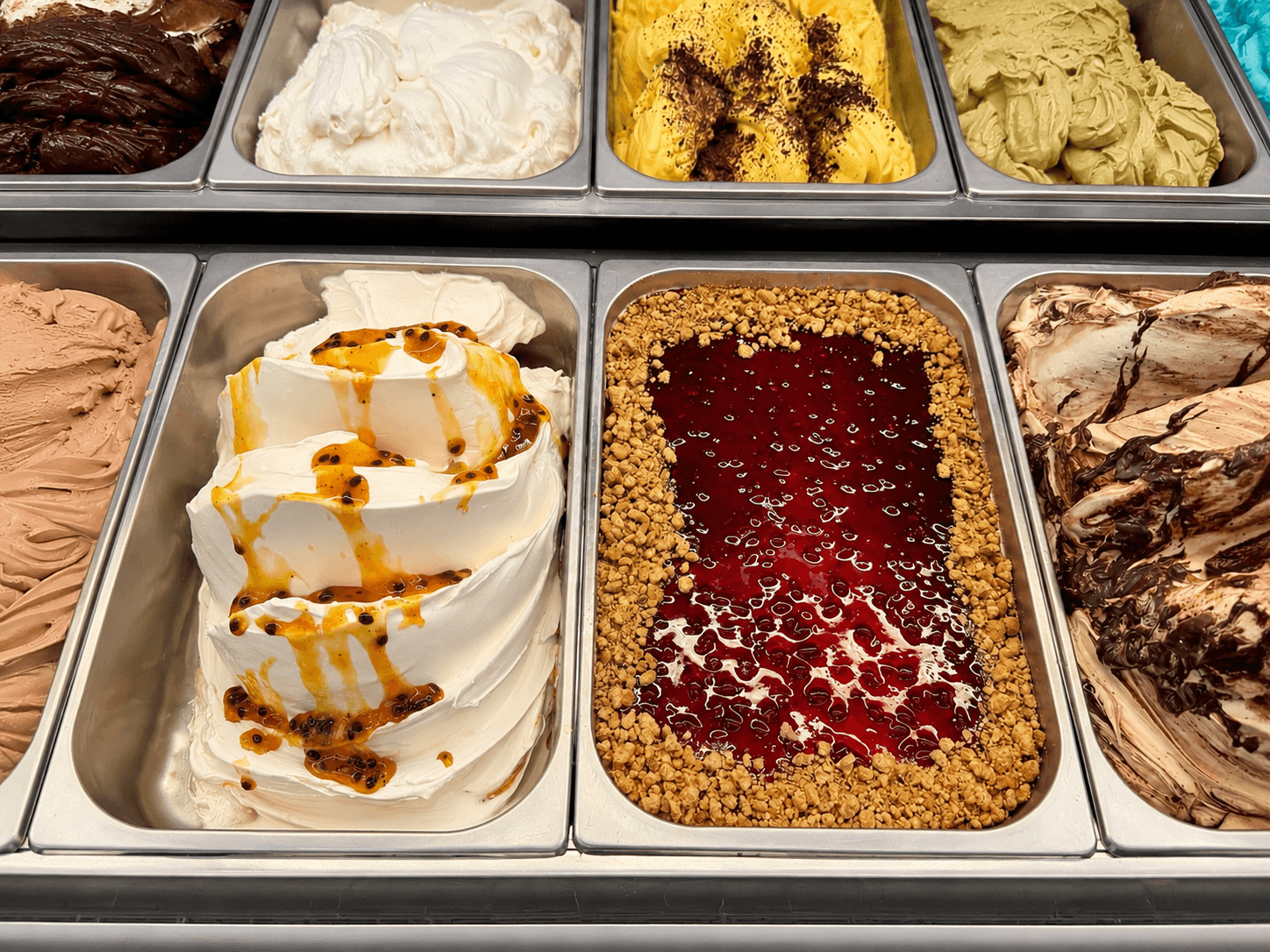 Bunte Auswahl an Eissorten in Metallbehältern, darunter Vanille mit Maracuja und Beerensoße mit Streuseln.