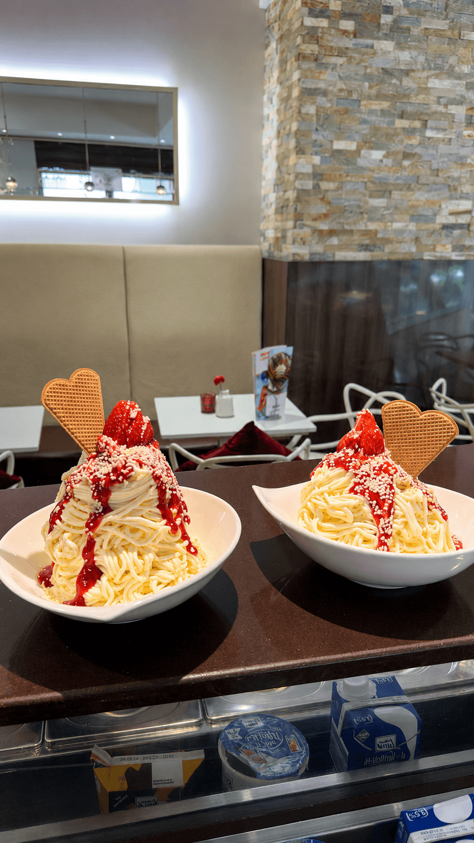 Zwei Schalen Spaghetti-Eis mit Erdbeersauce, weißen Schokoraspeln und herzförmigen Waffeln auf einem Tresen.