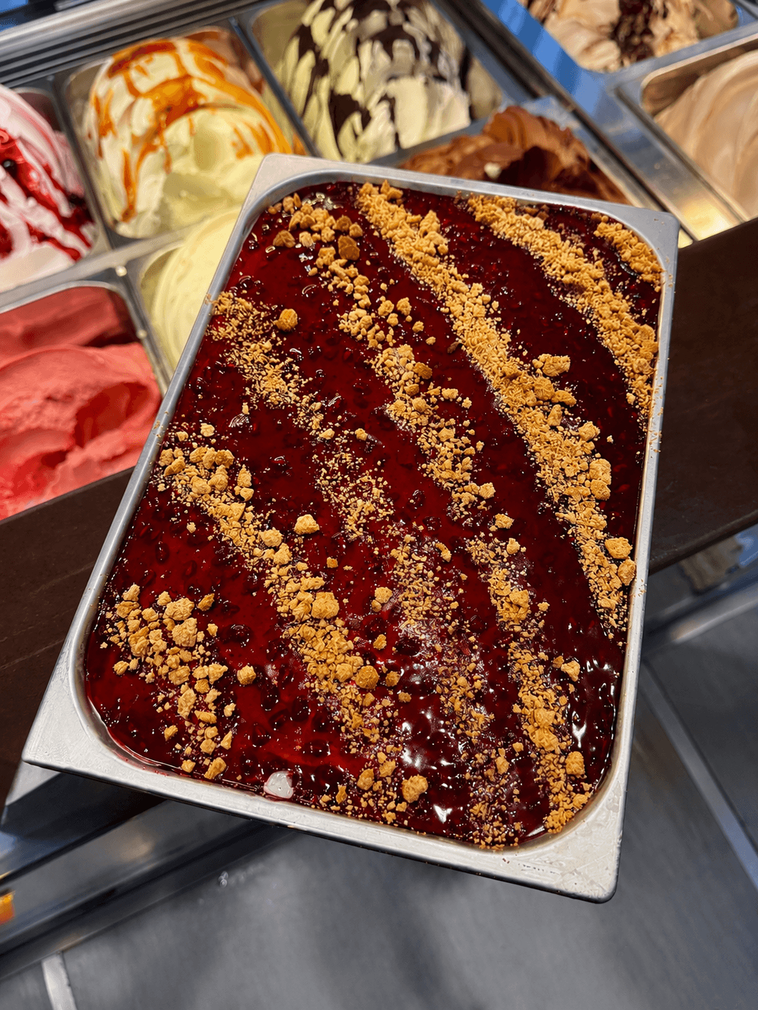 Dunkelrotes Beeren-Gelato in einer Metallschale, garniert mit diagonalen Streifen aus goldenen Kekskrümeln.