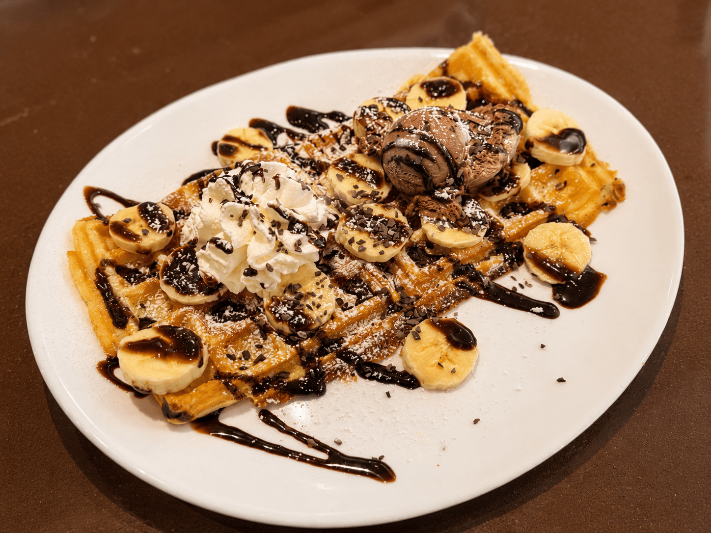 Goldbraune Waffel mit Bananen, Schokoladensauce, Schlagsahne, Schokoladeneis und Schokostreuseln auf einem weißen Teller.