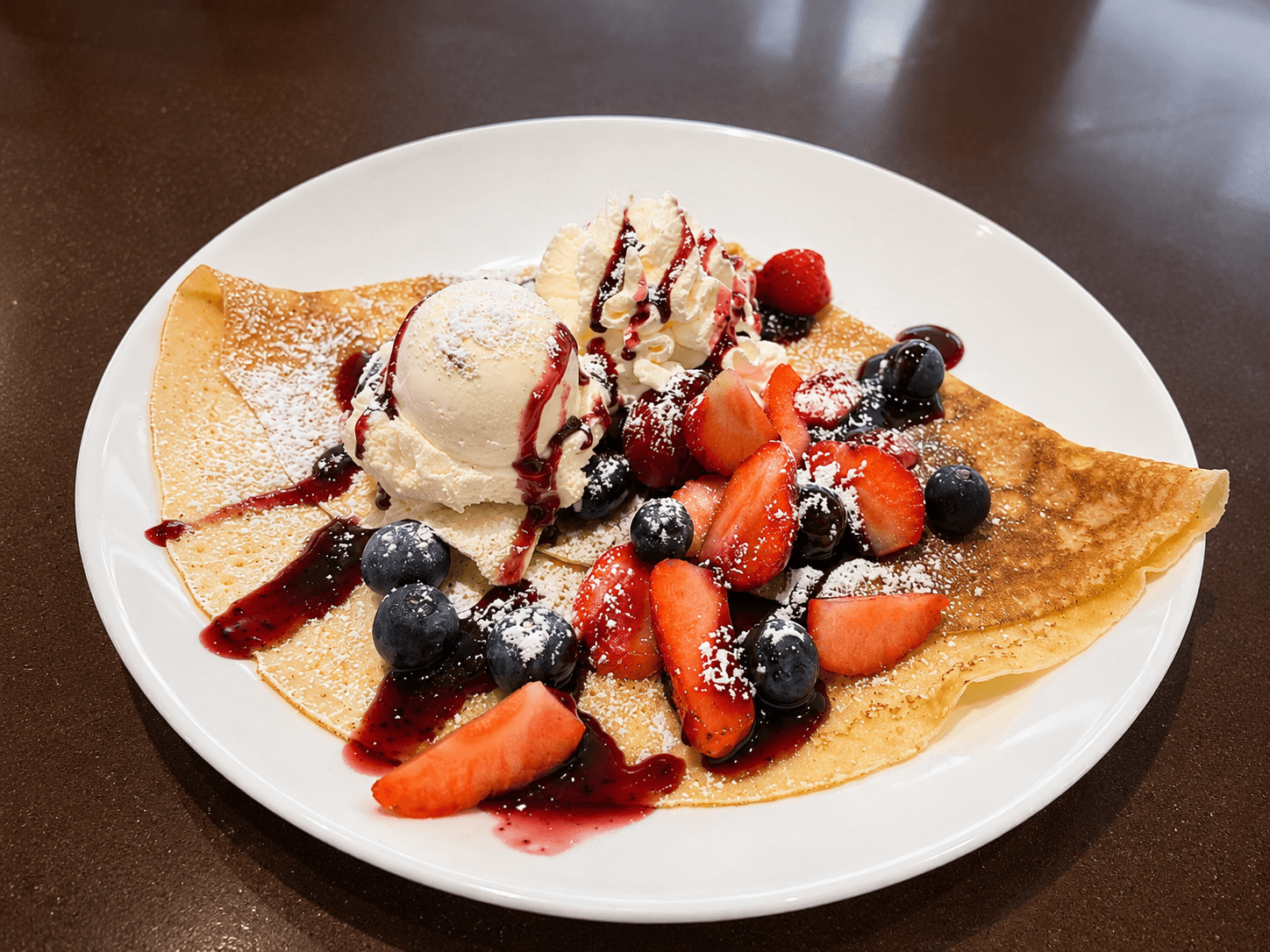 Goldbrauner Crêpe mit Vanilleeis, Schlagsahne, frischen Erdbeeren, Blaubeeren, Beerensauce und Puderzucker auf weißem Teller.