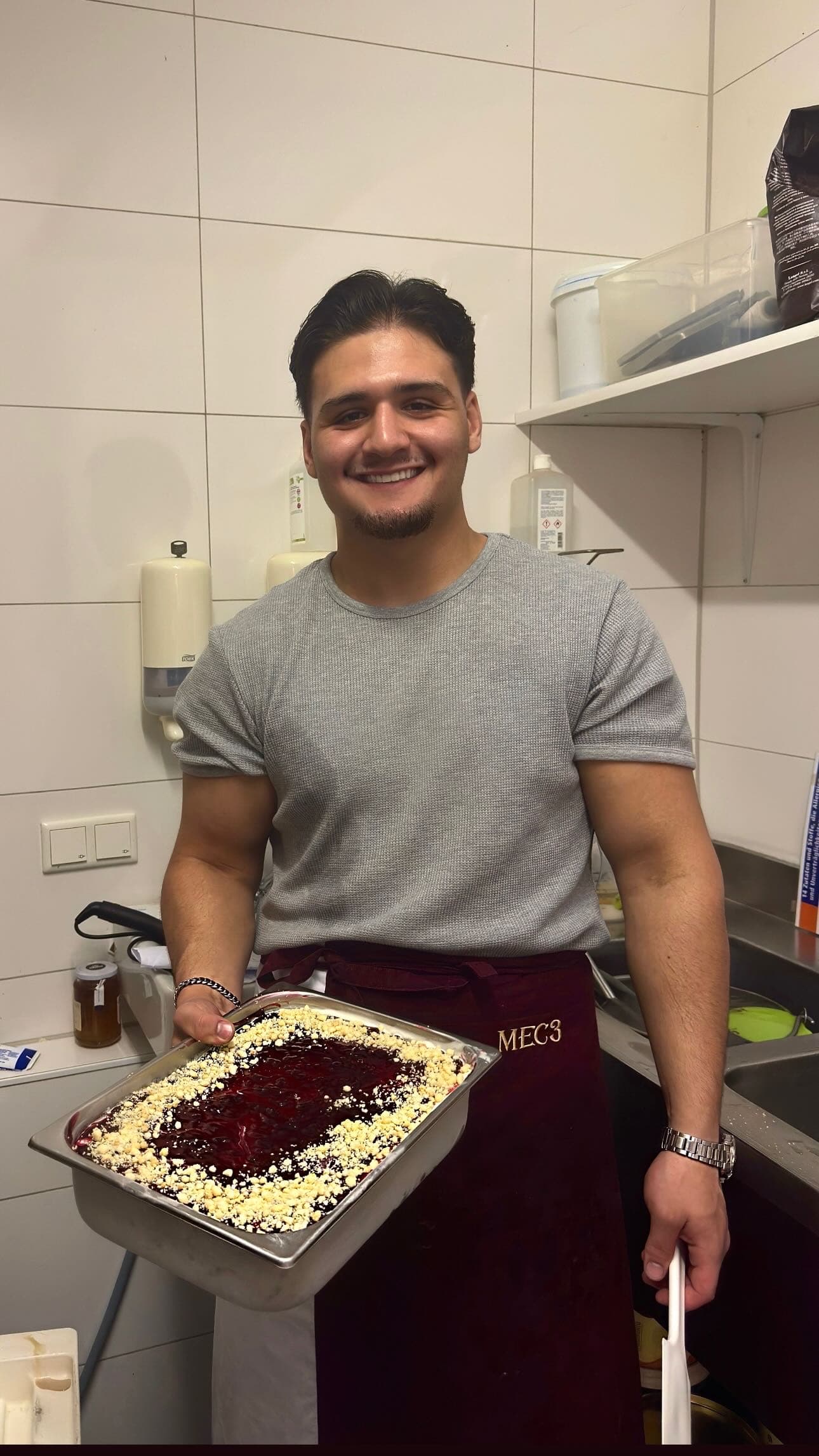 Ein lächelnder Mann in Schürze präsentiert ein Blech mit Beeren-Dessert in einer Küche.