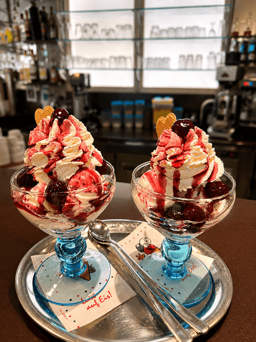 Zwei Eisbecher mit Schlagsahne, Kirschsauce und Kirschen auf einem silbernen Tablett in einem Eiscafé.