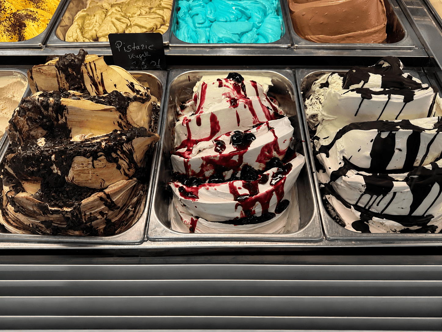 Bunte Eissorten in Metallbehältern, dekoriert mit Schokoladen- und Fruchtsaucen, präsentiert in einer gläsernen Eisvitrine.