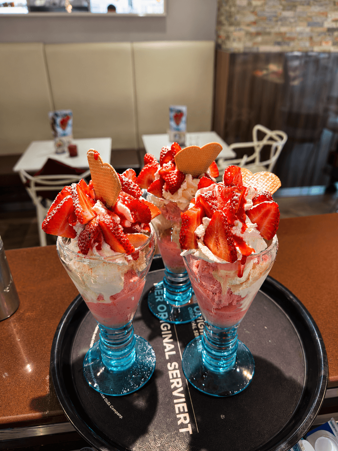 Drei üppige Erdbeereisbecher mit Schlagsahne, frischen Erdbeeren und Waffelherzen in blauen Gläsern auf einem Tablett.